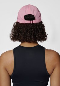 Rosa keps med justerbar rem, i bomullsmaterial, rund krona och broderad logotypdetalj, sedd från baksidan ovanpå lockigt hår.