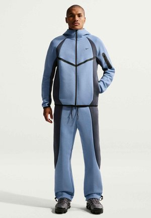 Mann steht in einem blauen und dunkelgrauen Nike-Trainingsanzug mit Reißverschlussjacke, passenden Hosen und schwarzen Sportschuhen vor einfarbigem Hintergrund.