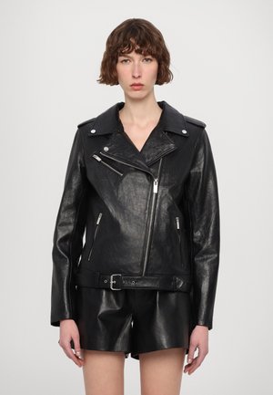 Schwarze Leder-Bikerjacke mit Reverskragen, silbernen Beschlägen, Reißverschlusstaschen und tailliertem Gürtel, kombiniert mit passenden Leder-Shorts.