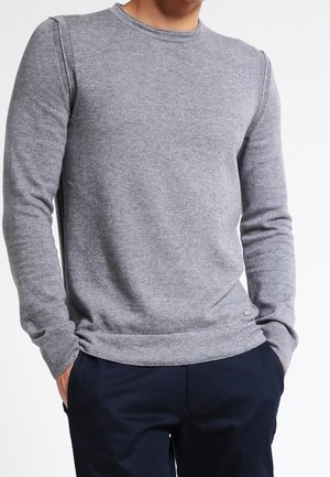 Mężczyzna ubrany w prosty szary sweter z długim rękawem, z rękami w kieszeniach, w połączeniu z ciemnoturkusowymi spodniami na białym tle.