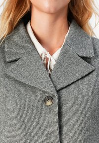 Cappotto grigio a spina di pesce con ampio colletto, chiusura a un bottone e tessuto testurizzato, indossato sopra una blusa bianca con scollatura a nodo.