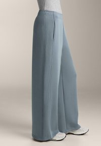 Svetlo modré nohavice s širokými nohavicami, hladkou textúrou, elastickým pásom a bočnými vreckami, skombinované so šedými teniskami.