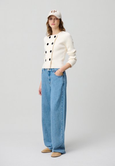 Maglione bianco a coste con pois neri, abbinato a pantaloni a vita alta in denim azzurro chiaro e scarpe slip-on beige.