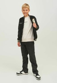 Giacca varsity nera con strisce bianche, felpa grigia, jeans scuri e sneakers nere con dettagli bianchi. Il modello si trova su uno sfondo neutro.