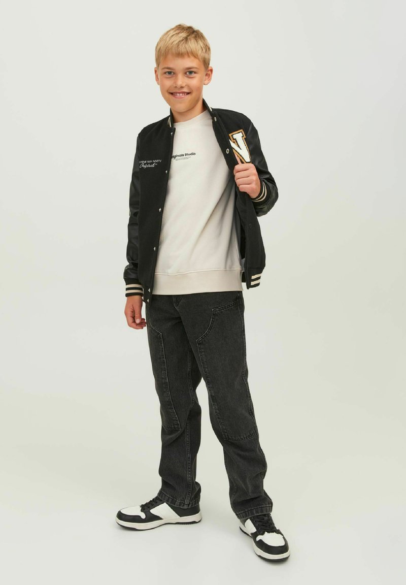 Giacca varsity nera con strisce bianche, felpa grigia, jeans scuri e sneakers nere con dettagli bianchi. Il modello si trova su uno sfondo neutro.