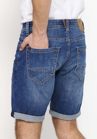 Shorts de mezclilla en un lavado azul medio, con dobladillos enrollados, bolsillos traseros y detalles de costura visibles. El material parece resistente y texturizado.