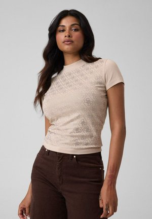 Femme aux longs cheveux foncés porte un haut beige à manches courtes à motifs et un pantalon brun foncé, debout devant un fond clair uni.