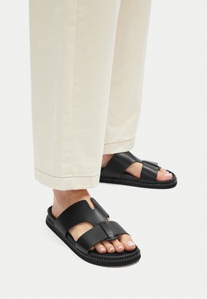 Čierne kožené sandále so širokými popruhmi a vykrojenými detailami. Hrubá podrážka s kontrastným stehom. Nosené s svetlými, voľne strihanými nohavicami.