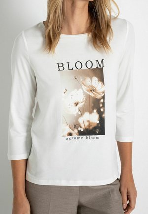 Kobieta w białej koszulce z rękawem do połowy przedramienia, z kwiatowym nadrukiem i tekstem "BLOOM" oraz "autumn bloom" z przodu.