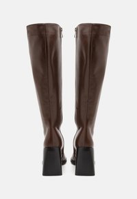 Bottes en cuir marron montant jusqu'aux genoux, avec une texture lisse, un talon carré et une fermeture éclair latérale. Construction robuste et silhouette élégante.