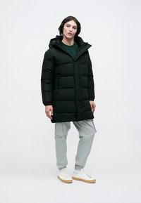 Abrigo acolchado negro con capucha, que presenta un cierre de cremallera y grandes secciones acolchadas. Llevado con pantalones cargo gris claro y zapatillas blancas.