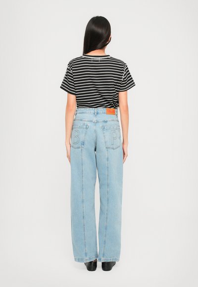 Woman with long dark hair wearing black and white striped t-shirt and high-waisted light blue jeans with letters "B" and "P" on back pockets.