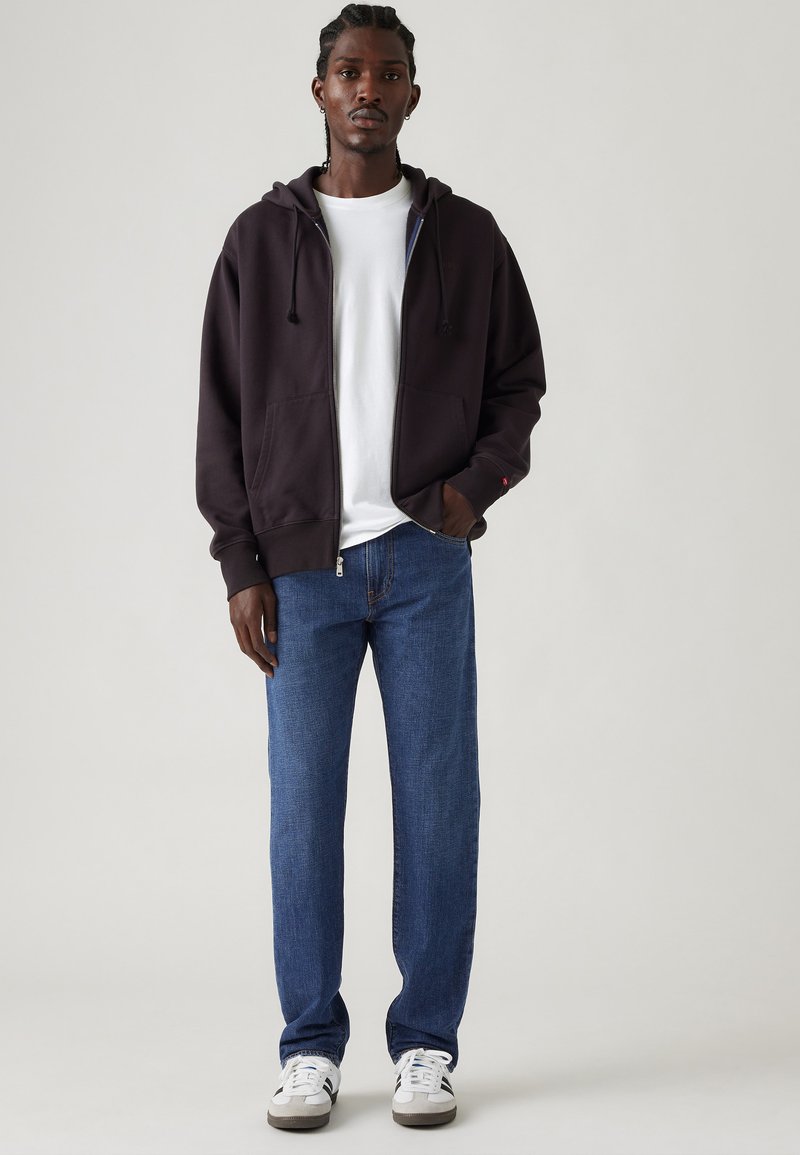 Ein Mann steht in einem schwarzen Zip-Hoodie, weißem T-Shirt, blauen Jeans und weißen Sneakern vor einem schlichten hellen Hintergrund.