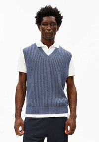 Blauw gebreid vest met verticale ribbels, V-hals, gedragen over een wit poloshirt. Heeft een losse pasvorm en zijsplitten voor comfort.