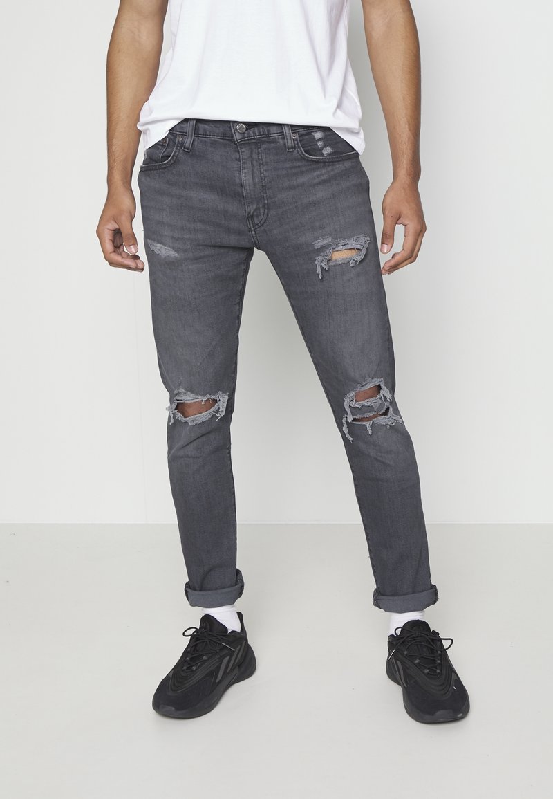 Blekna mörkgrå rivna jeans med flera slitna områden, slim fit design och uppvridna manschetter, som bärs tillsammans med svarta sportskor.