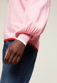 Ljusrosa tröja med puffärmar, försedda med en röd fåll och vita manschettdetaljer. Bärs med blå denimjeans.