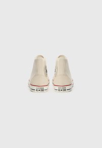 Beige Canvas High-Top Sneakers mit einem runden Logo-Patch und roten sowie blauen Streifen am Sohlenrand. Glatte Textur, klassisches Design.