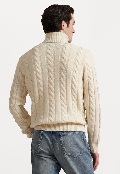 Crème kabelgebreide trui met hoge kraag, voorzien van textuurpatronen en geribbelde zoom, gecombineerd met lichtblauwe jeans.
