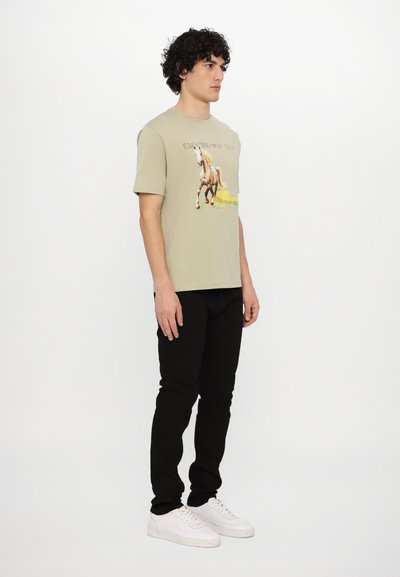 T-shirt vert olive avec un motif de cheval. Associée à un pantalon noir et des baskets blanches. Le modèle se tient devant un fond blanc uni.