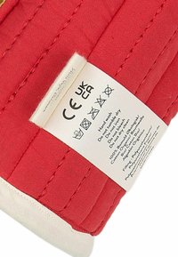 Eticheta din țesătură roșie cu cusături albe, care afișează instrucțiuni de îngrijire și detalii despre material. Textul include certificările CE și UKCA.