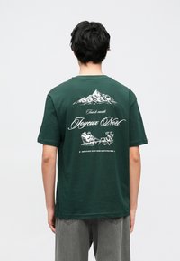 Camiseta de algodón verde con un gráfico blanco de montaña y el texto "Joyeux Noël" en la parte trasera, con un corte holgado y mangas cortas.