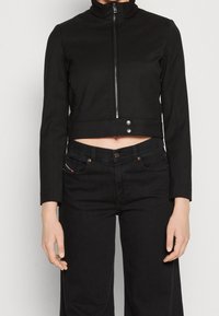 Svart kort jacka med hög krage, dragkedjestängning och två framknappar. Matchad med mörka jeans som framhäver en figurnära siluett.