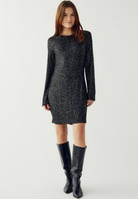 O femeie îmbrăcată într-o rochie mini neagră cu paiete și mâneci lungi, purtând cizme negre din piele până la genunchi, stând într-un fond alb simplu.