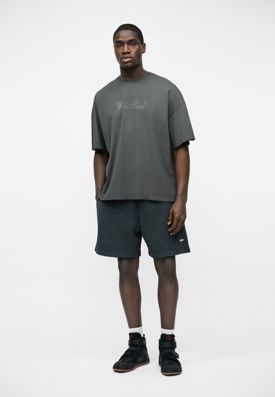 Jeune homme portant un t-shirt gris foncé surdimensionné, un short noir, des chaussettes blanches et des baskets montantes noires, debout devant un fond blanc uni.
