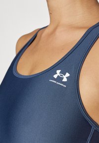 Temno modra športna majica brez rokavov z rebrasto strukturo in širokimi naramnicami. V zgornjem desnem kotu se nahaja bel logotip Under Armour.