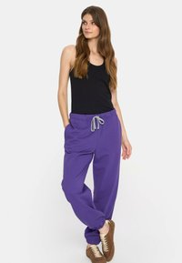 Kobieta w czarnym bezrękawniku, fioletowych spodniach dresowych na sznurki i brązowych trampkach, stojąca z jedną nogą skrzyżowaną na drugiej.