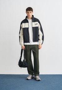 Αντιανεμικό μπουφάν της Nike σε μαύρο και κρεμ, με φερμουάρ στο μπροστινό μέρος και λογότυπο. Συνδυάζεται με λευκό μπλουζάκι, χακί παντελόνι και γκρι αθλητικά παπούτσια.