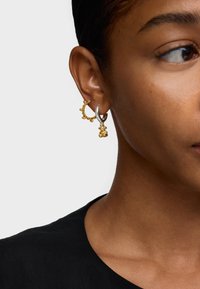 Złote i srebrne kolczyki z teksturowanym wzorem koła z okrągłymi akcentami, uzupełnione wiszącą ozdobą. Prosty i nowoczesny styl.