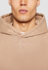 Béžova kapuca iz teksturiranega blaga; ima vrvico za nastavljanje kapuce in roza vezeno logo na prsih. Gladek zaključek, sproščen design.