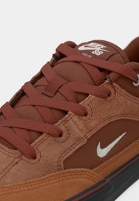 Sapatilha castanha em materiais de camurça e malha, com atacadores proeminentes e um logotipo branco da Nike na lateral. Detalhes texturizados por todo o lado.