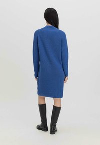 Stickad blå klänning med långa ärmar och ribbad textur, som når till mitten av låret. Kombinerad med knähöga svarta stövlar. Enkel, minimalistisk design.