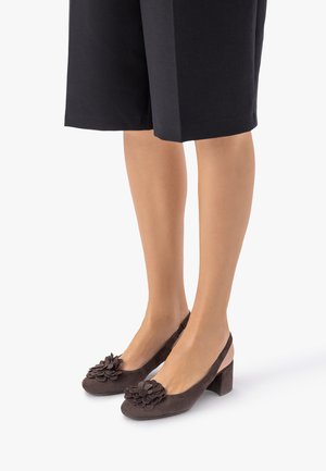 Žena oblečená v čiernych šortkách strednej dĺžky a hnedých semišových topánkach na hrubom podpätku s kvetinovými ozdobami na špičkách, stojaca na bielom pozadí.