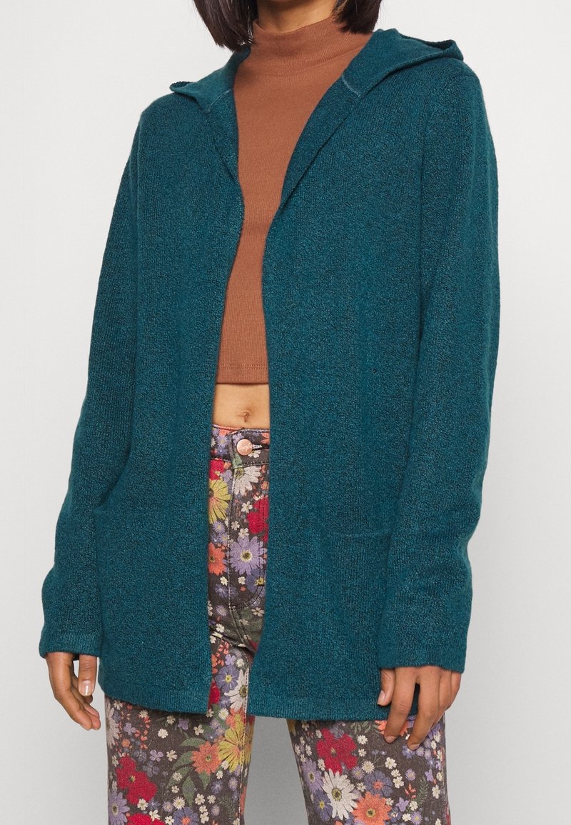 Teal huva cardigan i strukturerad stickning, med framfickor, öppen design, kombinerad med blommönstrade byxor, liknande klassisk avslappnad stil.