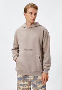 Sudadera con capucha en mezcla de algodón color topo con bolsillo frontal. Presenta el texto "CREE EN TI MISMO OCHENTA Y OCHO" centrado sobre el bolsillo.