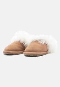 Egy pár világosbarna velúr papucs vastag fehér, bolyhos béléssel, slip-on stílusban, egy sima fehér háttér előtt kiemelve.