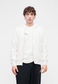 Ανοιχτό γκρι φούτερ με φερμουάρ και μπροστινές τσέπες, λευκή κοντομάνικη μπλούζα με στρογγυλή λαιμόκοψη και την επιγραφή "OFFICIAL", και σκούρο παντελόνι φόρμας με πλαϊνές τσέπες.