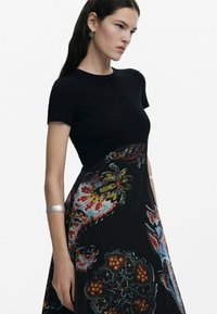 Zwarte geribbelde topjurk met een vloeiende rok met kleurrijke bloem- en abstracte patronen, aangevuld met een zilveren armband en oorbellen.