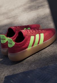 Pair of red Adidas sneakers with neon green stripes and heels, and semi-translucent gum soles resting on a gray surface with shadow.