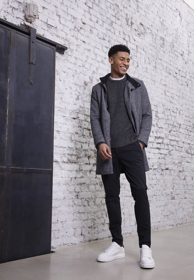 Abrigo gris texturizado sobre un suéter oscuro, combinado con pantalones de rayas negras y zapatillas blancas, con un fondo de pared de ladrillo.