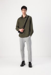 Olive green button-up shirt, grey tailored pants, and black lace-up shoes. The shirt has long sleeves and a relaxed fit, with a black shoulder bag.