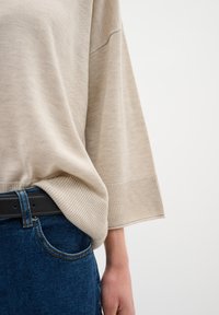 Beige, laza kötött pulóver széles ujjakkal, bordázott szegéllyel és sima textúrával, sötétkék farmernadrággal és fekete övvel párosítva.