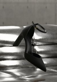 Μαύρο γυναικείο παπούτσι με ψηλό τακούνι και λουράκι στον αστράγαλο, κρεμασμένο πάνω από ασημί μεταλλικό καναπέ με κουμπιά, μπροστά σε γκρι τοίχο.