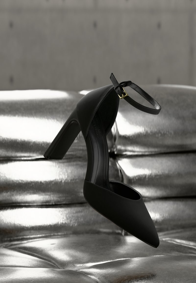 Chaussure à talon haut noire avec bride à la cheville suspendue au-dessus d'un canapé argenté métallique capitonné, devant un mur gris.