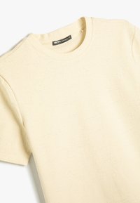 Camiseta de cuello redondo de manga corta en color beige claro, hecha de una tela texturizada, con un patrón sutil y una etiqueta en el cuello.