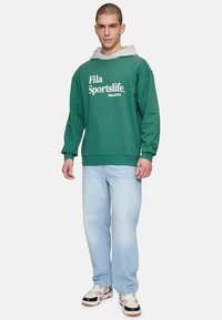 Felpa verde in pile con cappuccio grigio, caratterizzata da testo bianco "Fila Sportslife Since 1974". Indossata con jeans dritti blu chiaro e sneakers multicolori.