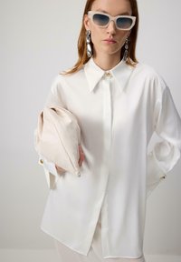 Biela oversized košeľa z hladkej látky s gombíkmi a zlatým akcentovým gombíkom. Obsahuje béžovú kabelku a šedé slnečné okuliare.